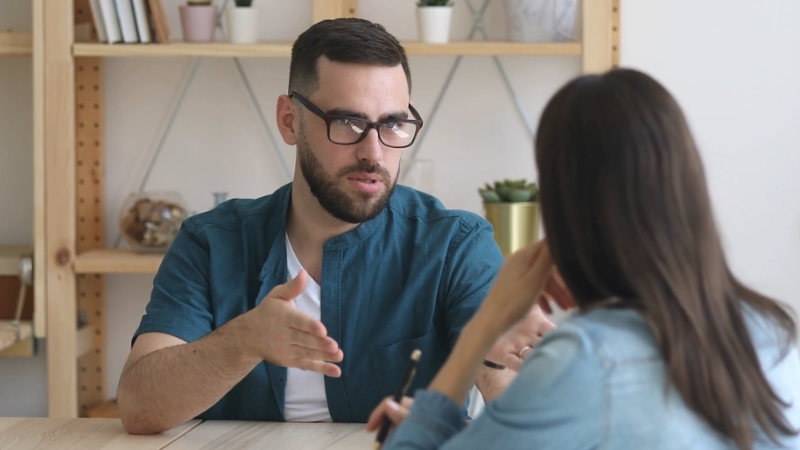A man with glasses and a beard speaks to a woman across a table