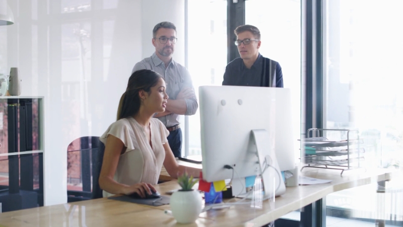 A woman discusses her performance at a desk while two male colleagues stand nearby