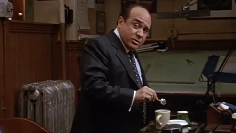 Lawrence Garfield stands by his desk, stirring coffee while making a point in a scene from Other People’s Money
