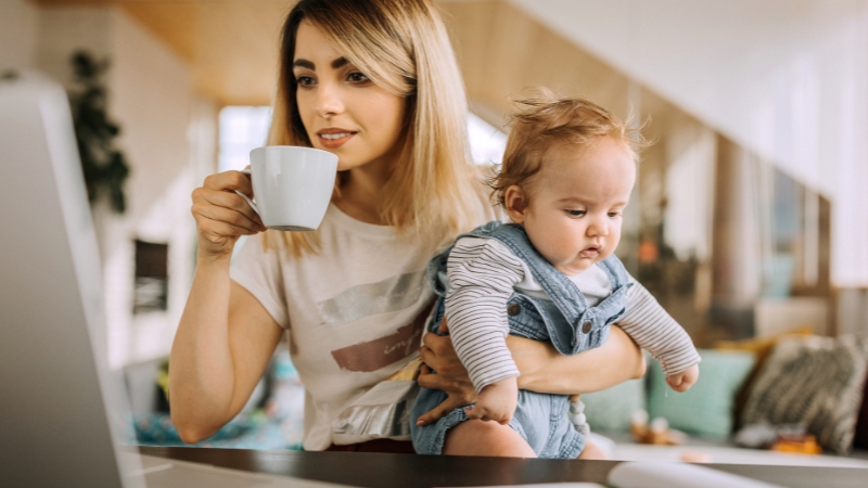 A woman holds her baby and a cup of coffee while working on a computer, managing her work-life balance at home