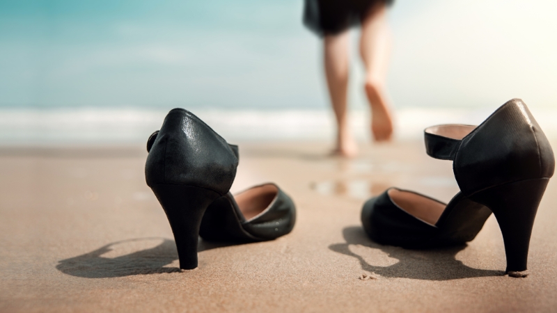 A woman walks barefoot on the beach, leaving her black heels behind as she takes time off to unwind