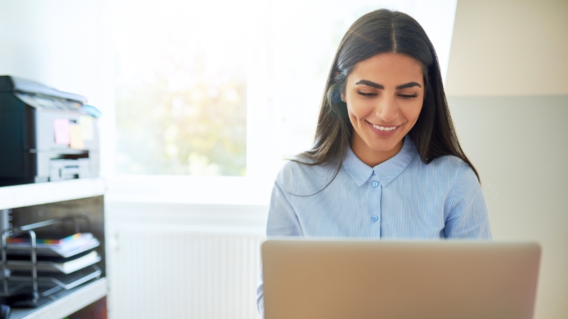 Young Gen Z professional working remotely with a smile, focused on her laptop in a bright home office