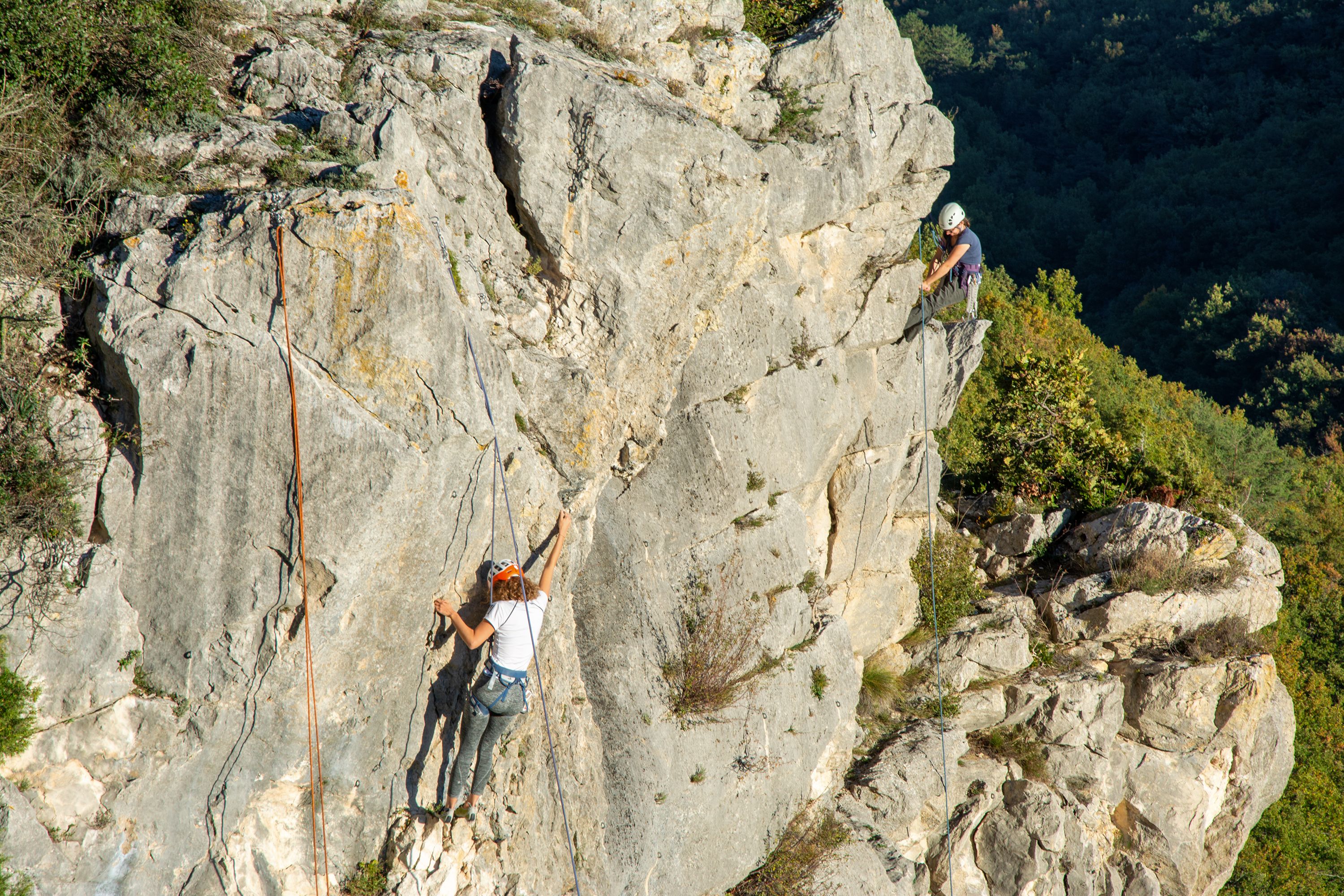 Two women climb a sheer outdoor rock face using ropes and safety gear.