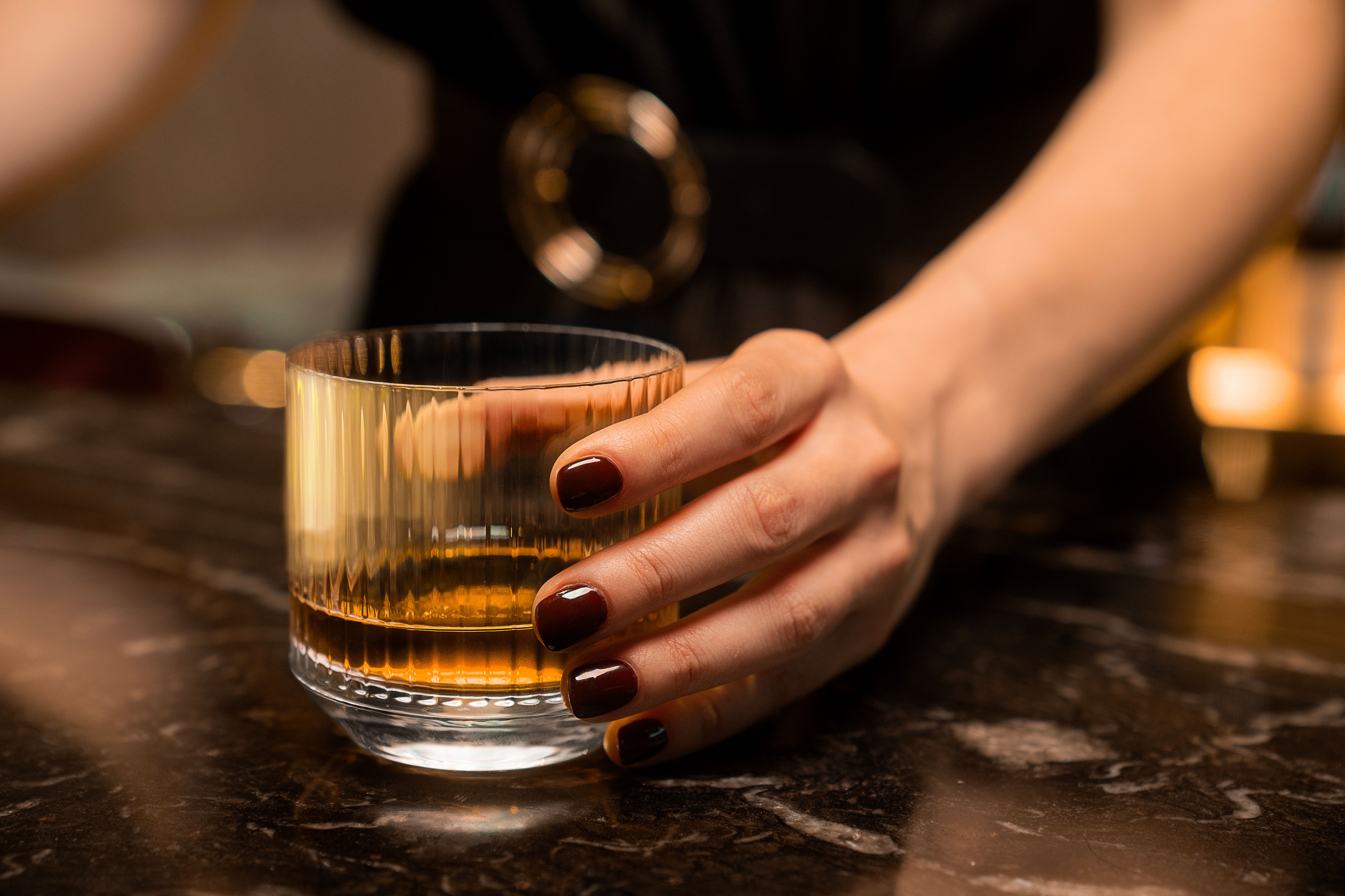 Woman's hand holds a glass of whiskey in a warmly lit setting, suggesting a whiskey tasting experience.